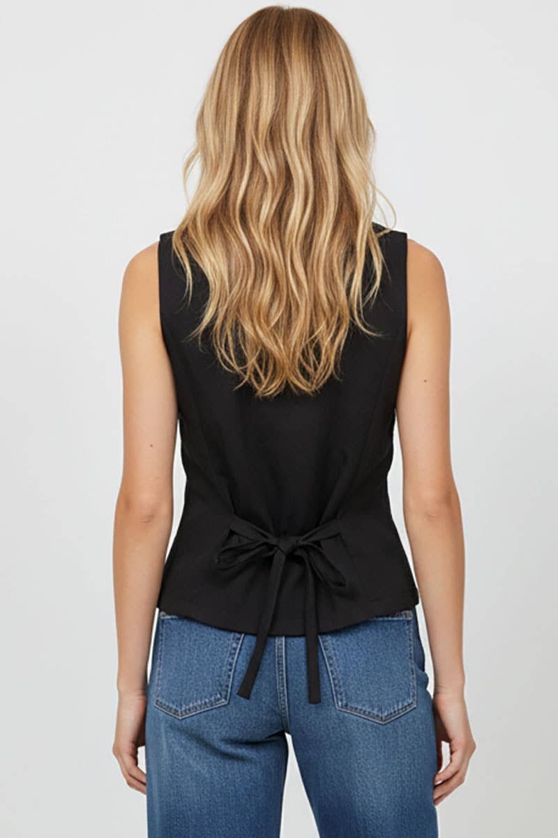 Person wearing a black sleeveless top with a tie detail and blue jeans on a plain background