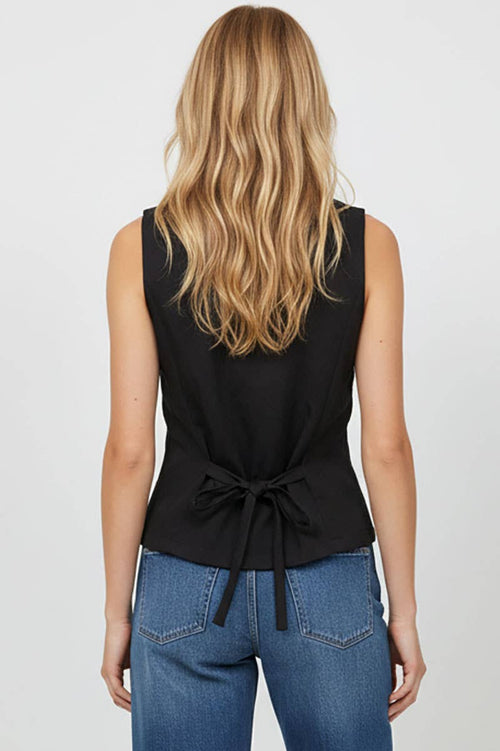 Person wearing a black sleeveless top with a tie detail and blue jeans on a plain background